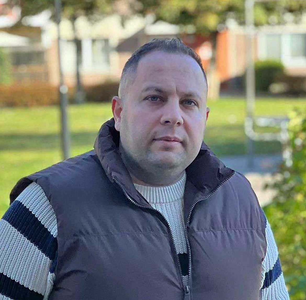 Arbetsledaren Yasser på Amalia Städ och inredning poserar utomhus i en blå väst och stickad tröja, med en bakgrund av grönska och bostadshus